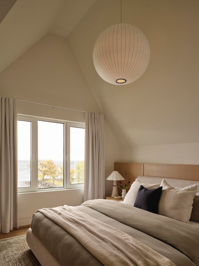 Bedroom with vaulted ceiling and spherical pendant light above a window overlooking the water