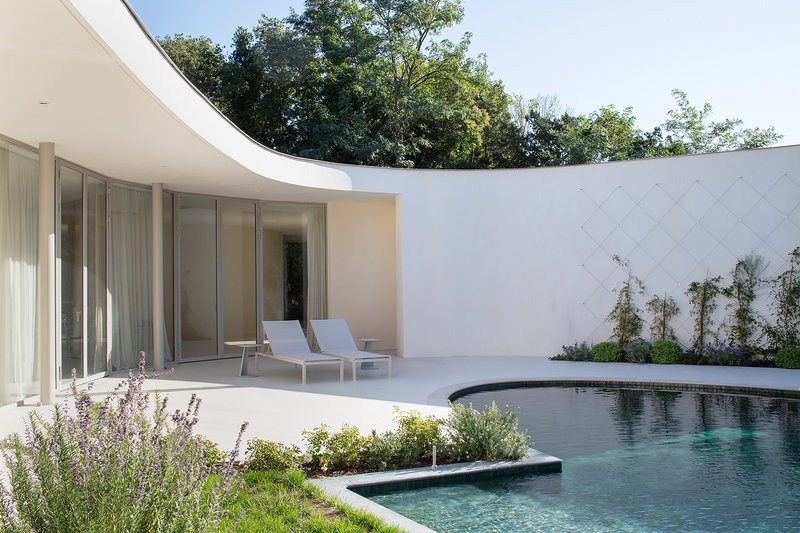 Pool terrace with glass pavilion, curved overhang, and planted beds along white tiled wall