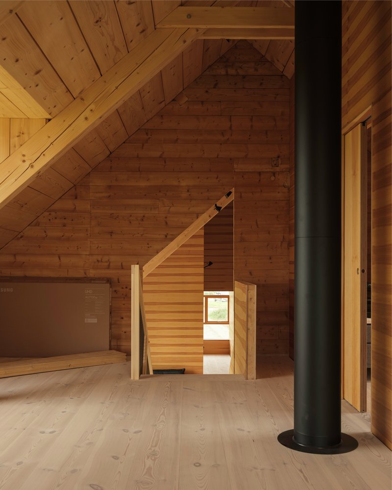 Timber interior with exposed diagonal ceiling planks, freestanding black flue pipe, and open stairwell beyond doorway