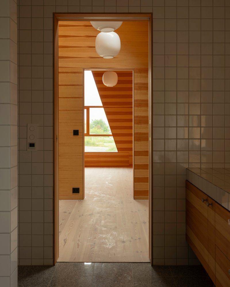 Doorway from white tiled bathroom into timber hallway with spherical pendant lamp and angled window framing greenery