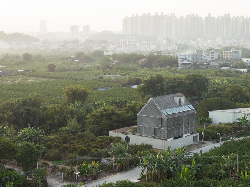 Elevated view of pitched-roof concrete house surrounded by agricultural fields and distant towers in morning haze