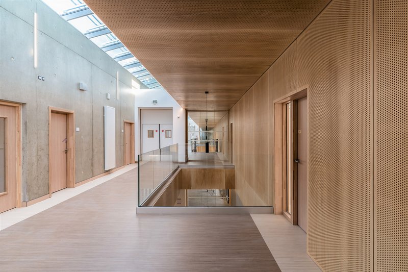 Glazed bridge opening spanning an interior void with perforated timber ceiling and exposed concrete walls