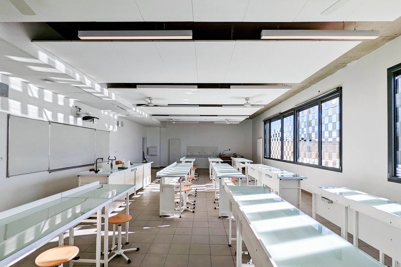 Laboratory classroom with white workbenches and stools beneath linear ceiling fixtures and suspended equipment