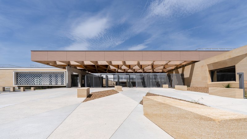 Entrance canopy supported by angled timber columns under a bright midday sky