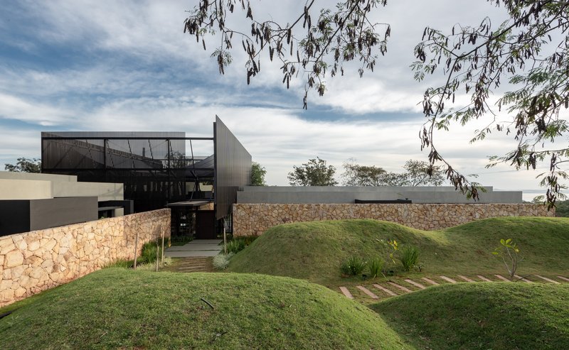 Entrance path between stone retaining walls and grassed berms beneath corrugated metal volumes and eucalyptus branches