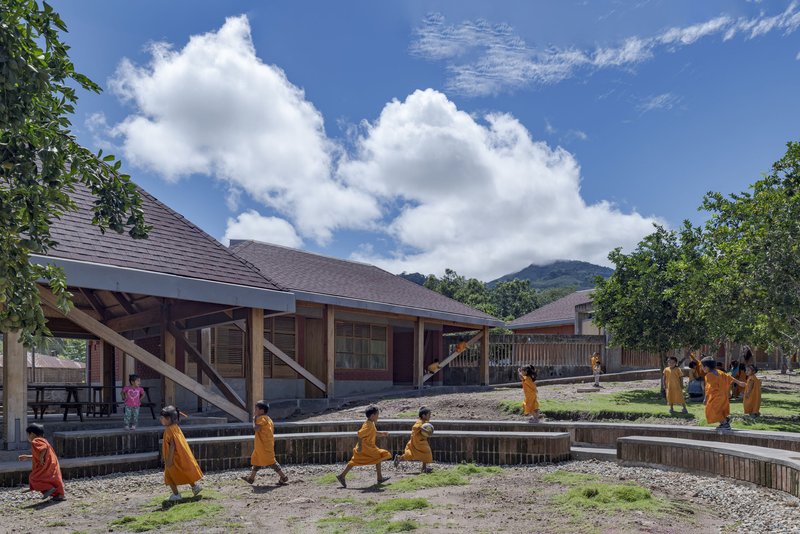 Courtyard with low-slung pitched roof buildings and stepped timber retaining walls as children walk along the pathways