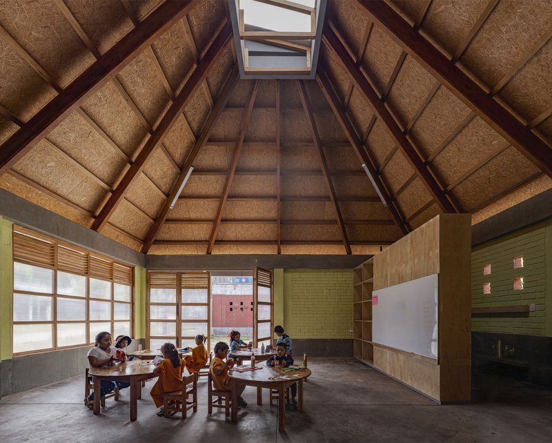 Interior classroom with pyramidal roof structure, exposed timber rafters, and children seated at low wooden tables