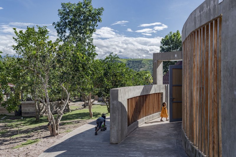 Elevated concrete walkway with vertical timber screens, children walking beside trees under a cloudy sky