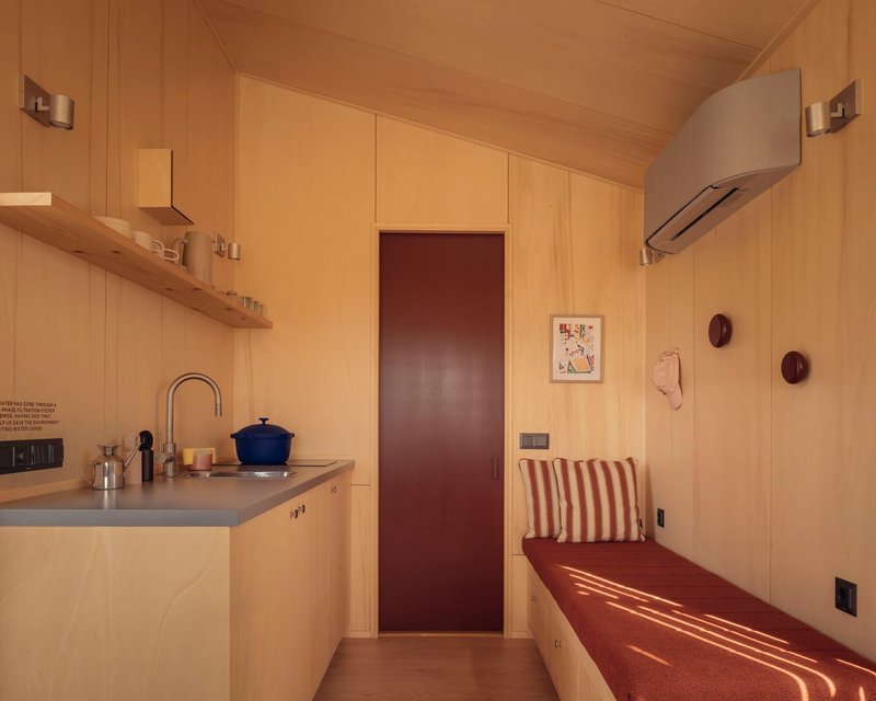 Compact kitchenette with birch plywood walls, dark red door and built-in upholstered bench in afternoon light