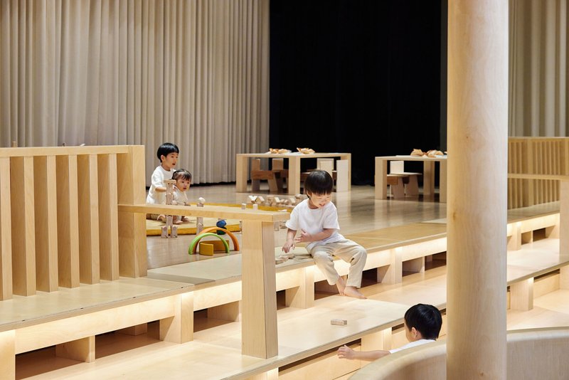 Interior play space with stepped light wood platforms and children sitting among fabric curtains