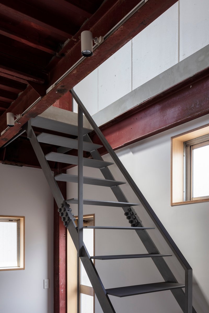 Steel stair rising past plywood-framed windows beneath the exposed timber ceiling structure