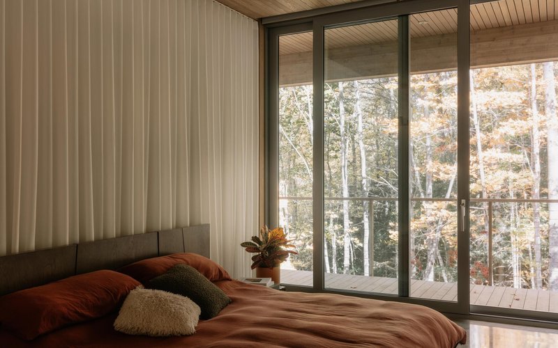 Bedroom with floor-to-ceiling glazing opening to a balcony overlooking autumn trees in soft morning light