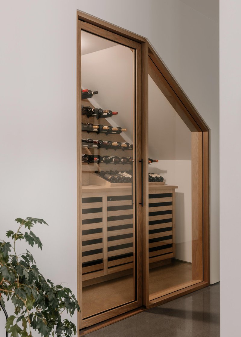 Angled glass door framing a wine cellar with horizontal slat cabinet and bottle racks beside a potted plant