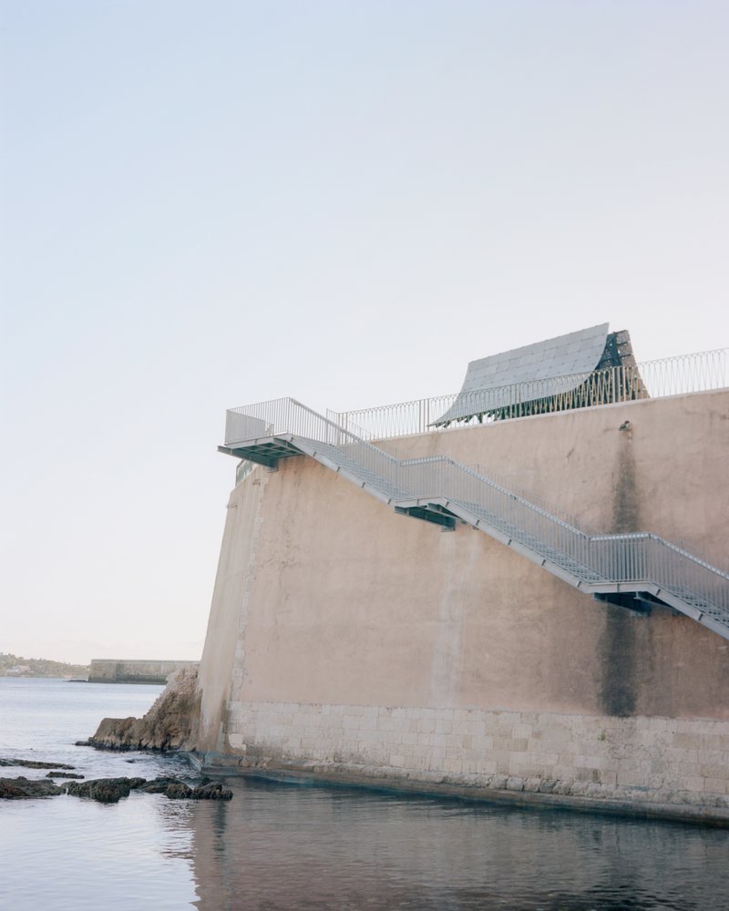 Metal staircase ascending the exterior concrete wall above rocky shoreline water