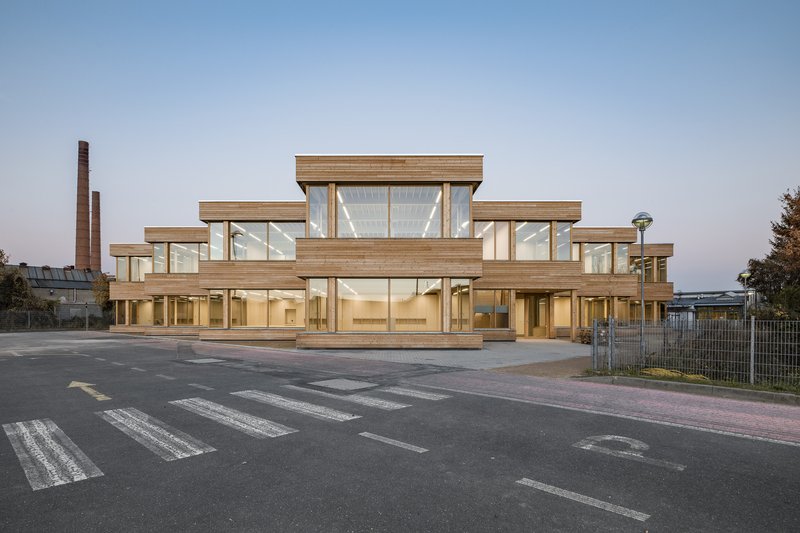 Stepped timber and glass facade fronting a crosswalk at dusk with brick chimneys beyond