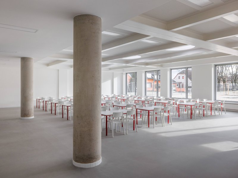 Interior dining hall with cylindrical concrete columns and rows of red and white chairs below a coffered ceiling
