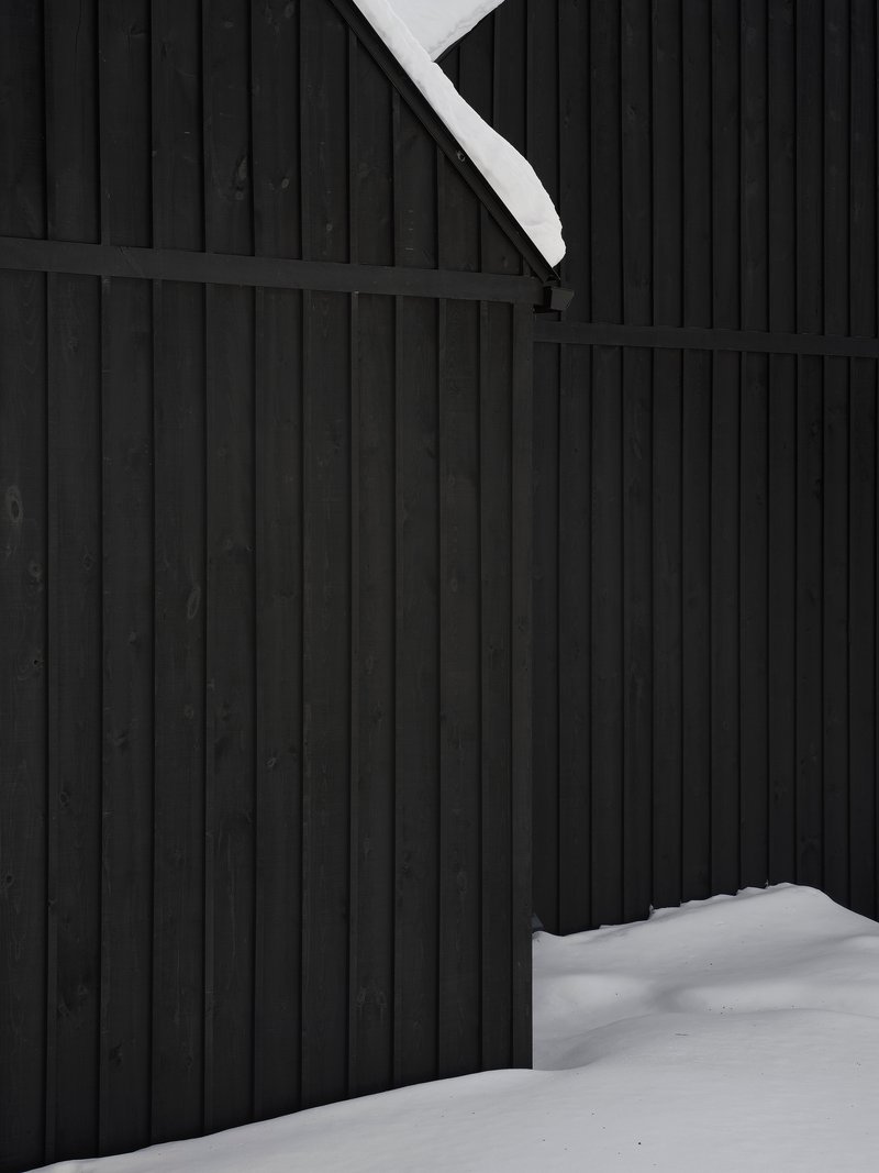 Close-up of black vertical wood siding meeting snow-covered roof edge and ground plane