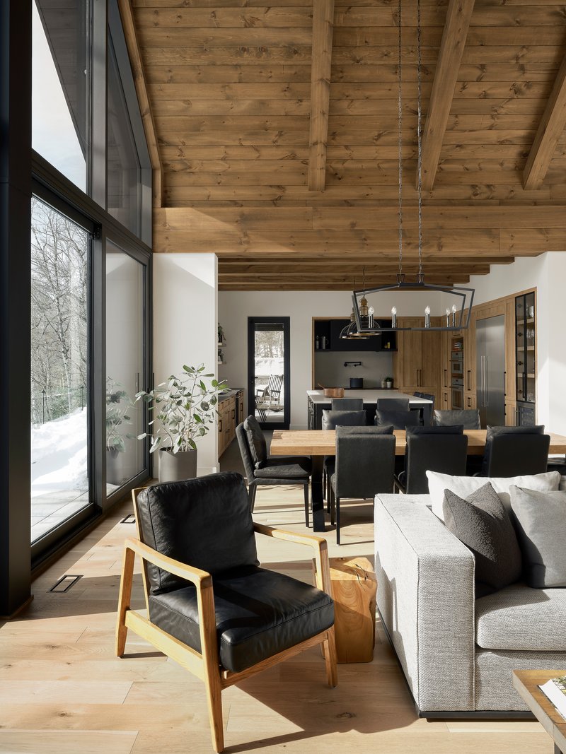 Open living space with exposed timber ceiling beams and tall glazing overlooking snowy forest