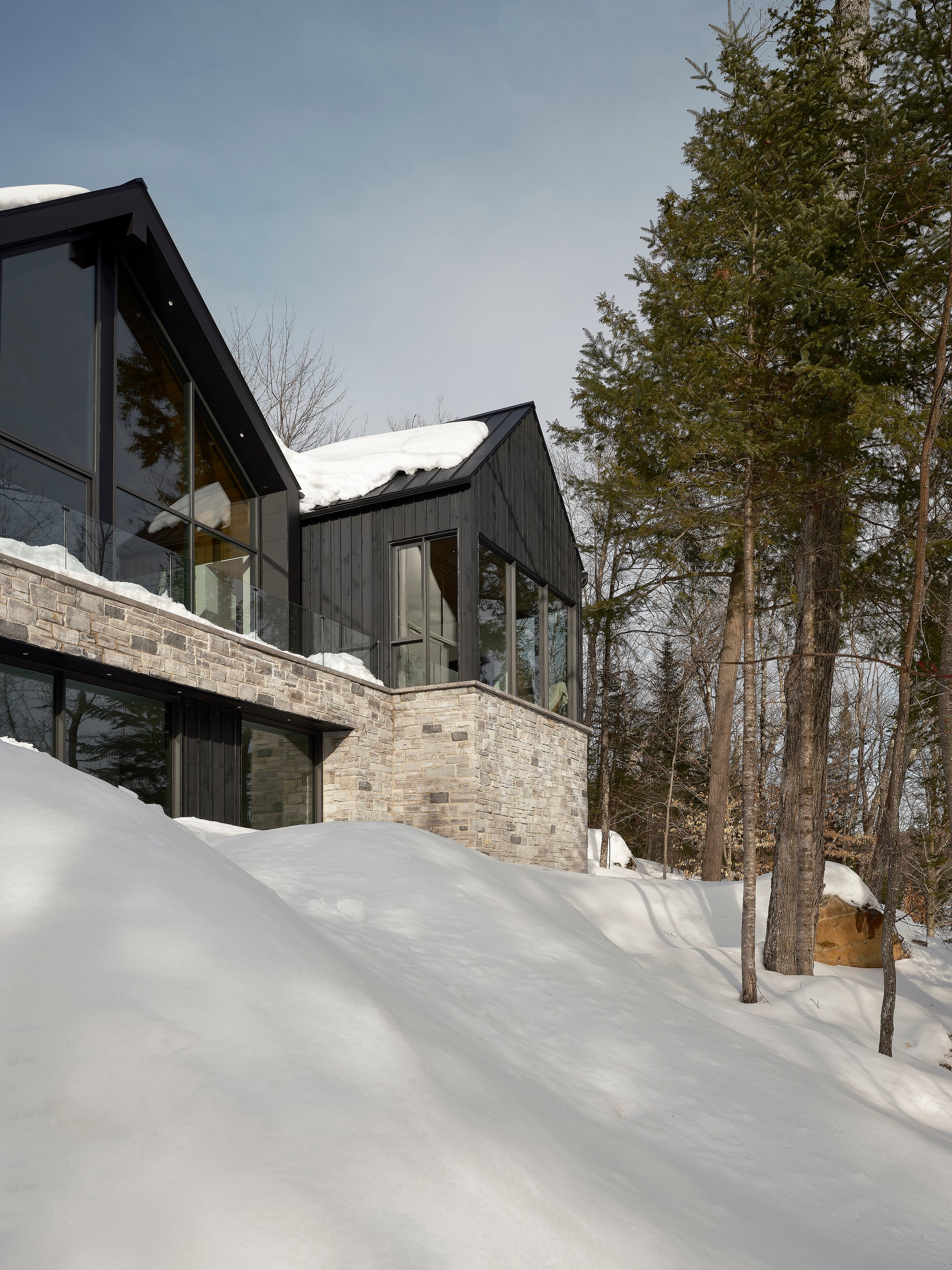 Nathalie Thibodeau Sets Three Dark Gabled Volumes on a Stone Plinth Between Road and Lake