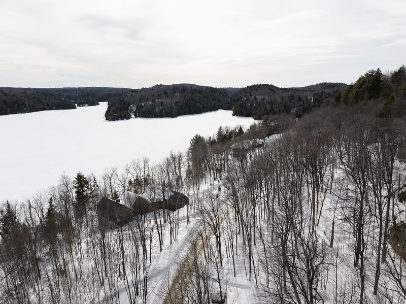 Aerial view of the dark pitched roof volumes nestled among bare winter trees beside a frozen lake