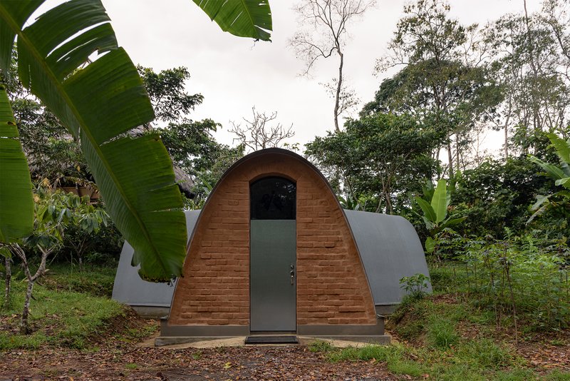 Front facade with timber arched door and grey metal vault roof set among banana plants and trees