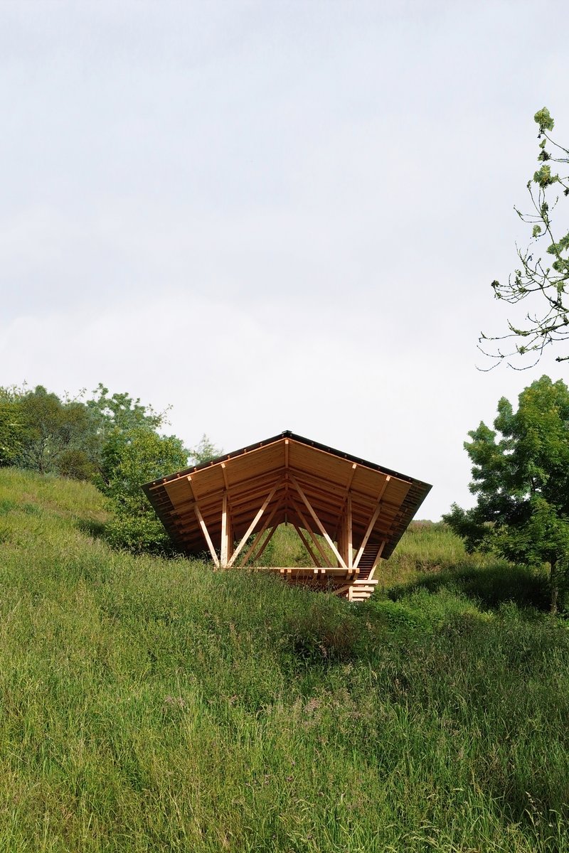 Timber pavilion with exposed triangular roof trusses set into a grassy hillside under overcast skies