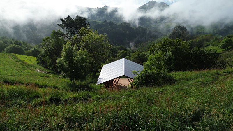 Small tilted pavilion with corrugated metal roof set into a green hillside with mist drifting through the trees