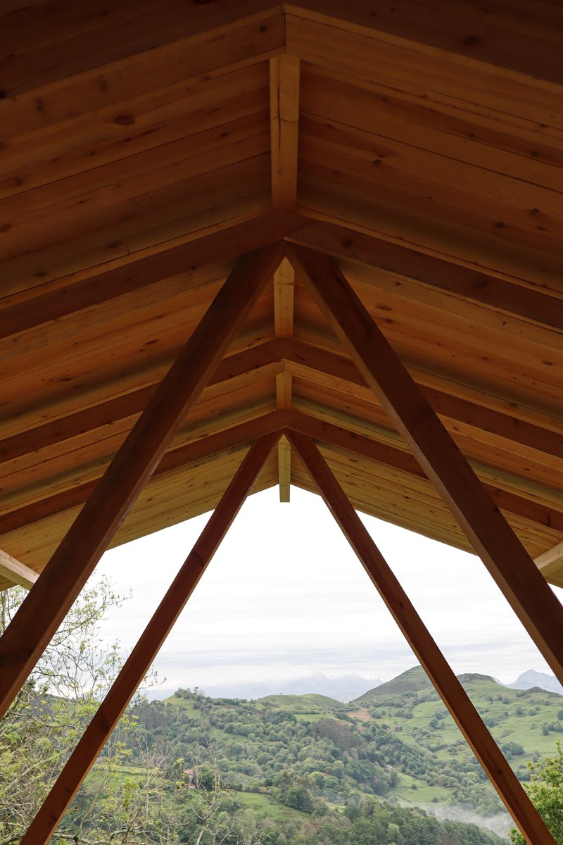 Triangular view through branching timber trusses and tongue-and-groove ceiling toward rolling green hills