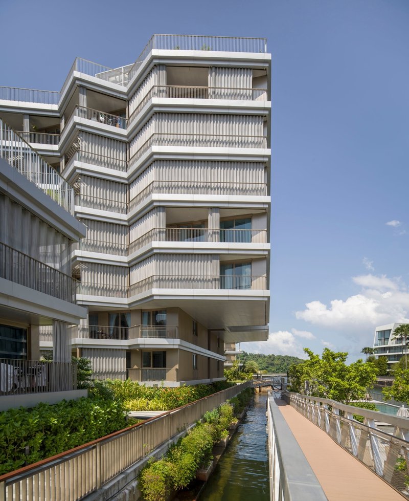Zigzagging facade with vertical louvers and recessed balconies above a planted canal under blue sky