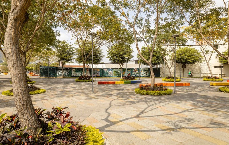 Paved plaza with circular planting beds and orange benches beneath dappled tree shadows