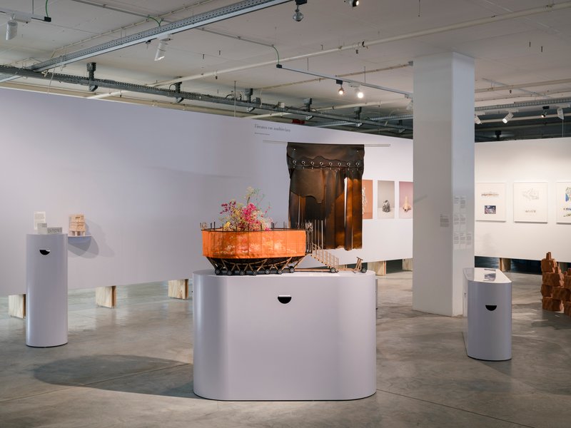 Exhibition space with sculptural installation on white plinth beneath exposed ceiling ducts and track lights