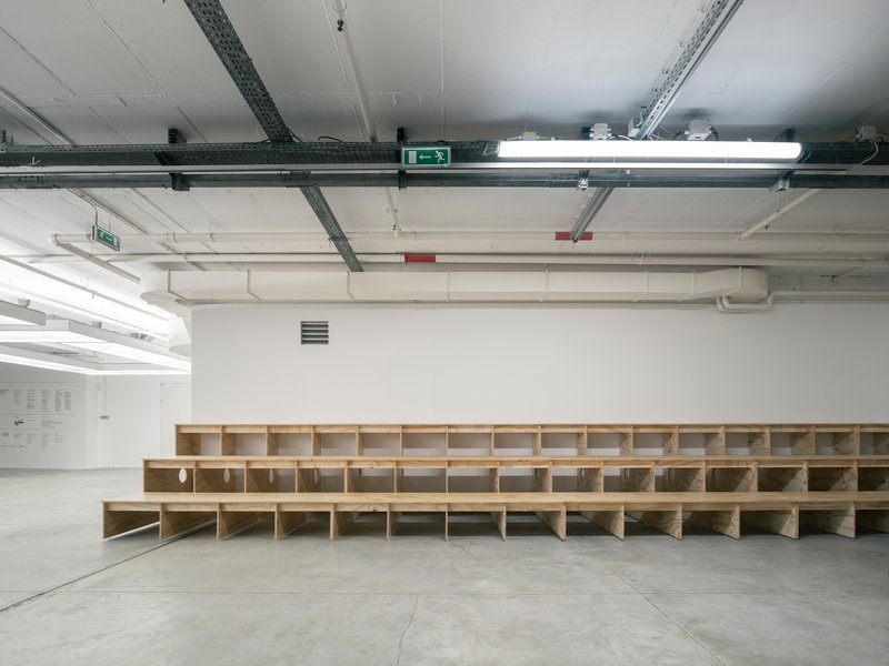 Long tiered plywood seating structure with stepped risers spanning the width of a white gallery space