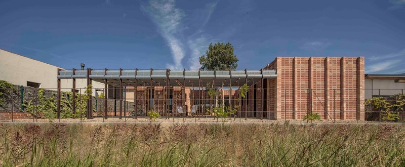 Street elevation showing perforated brick facade with timber pergola and native grasses in foreground under clear sky