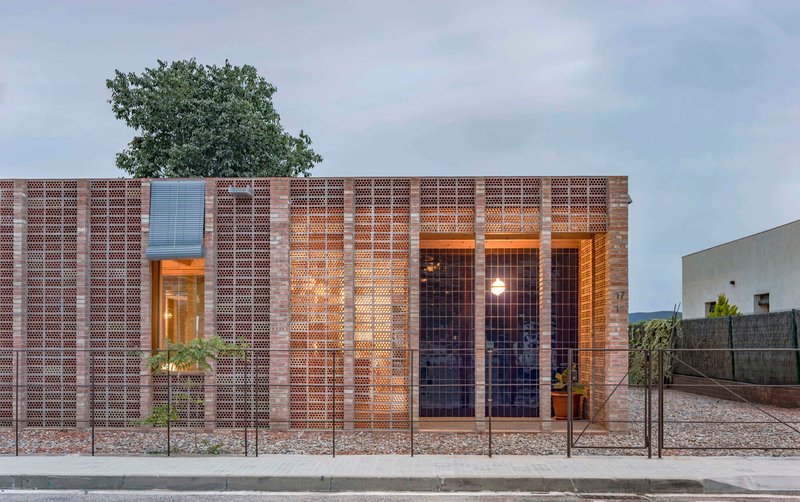 Street facade at dusk showing illuminated interior through perforated brick and steel mesh screens with tree above