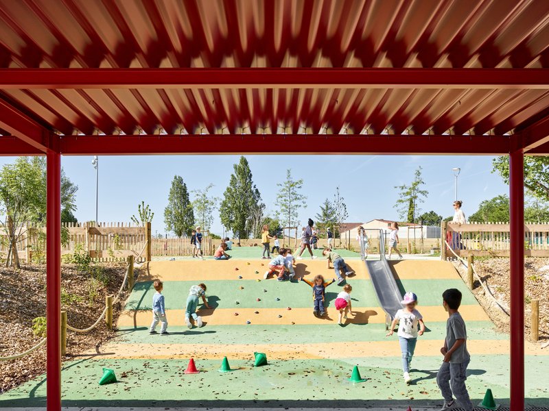 Red steel canopy framing a playground with children on a climbing hill and scattered play equipment