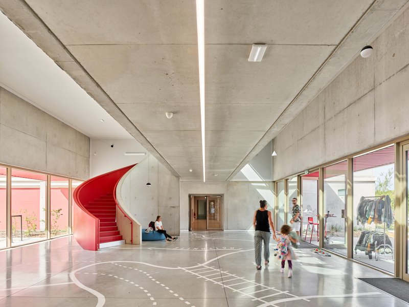 Concrete interior lobby with a red helical staircase and linear ceiling skylights illuminating the polished floor