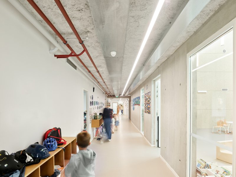 Hallway with exposed red conduit along the concrete ceiling as children walk past storage cubbies