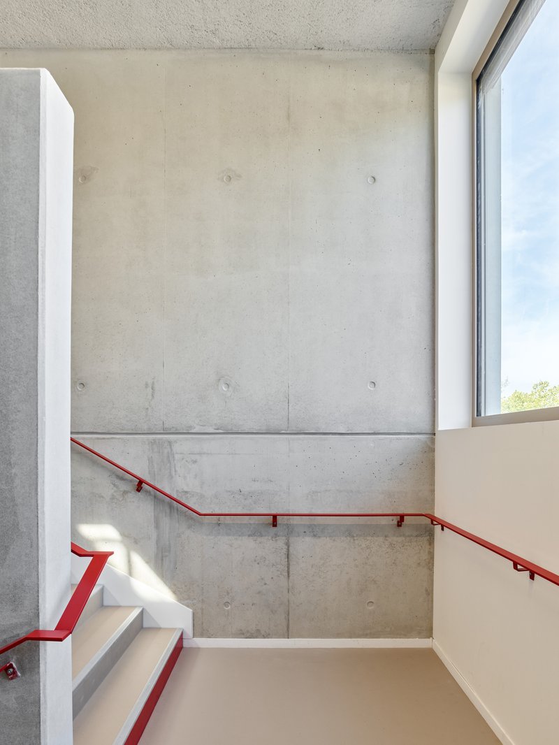 Stairwell with cast concrete walls, red steel handrail and natural light from a corner window