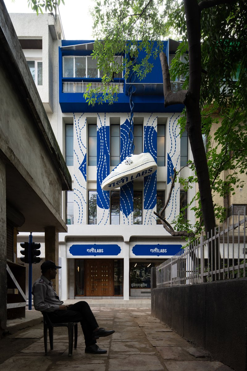 Street facade with blue and white painted mural behind overhanging trees and seated figure on paved walkway