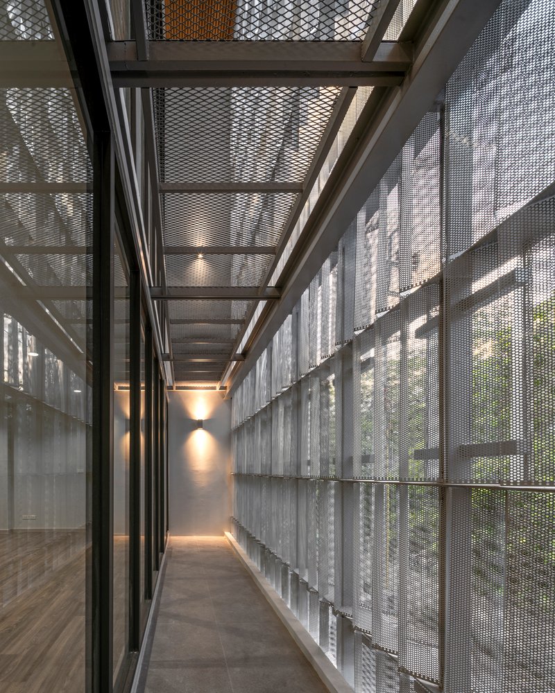 Corridor enclosed by perforated metal screens and exposed steel beams filtering dappled light from exterior planting