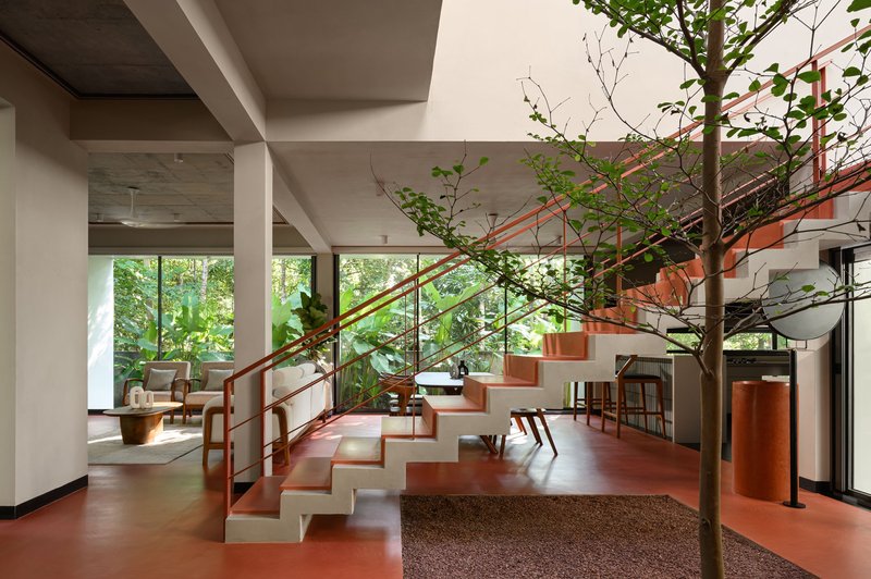 Interior staircase with red metal railings ascending past planted tree in terrazzo floor well
