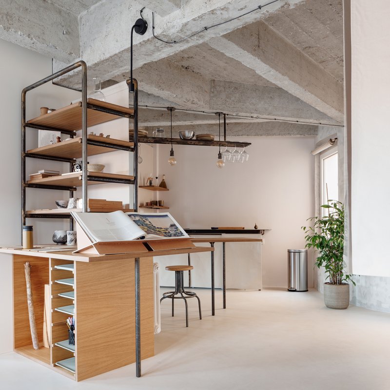 Tilted drafting table with modular oak storage unit and suspended steel shelving under concrete beams
