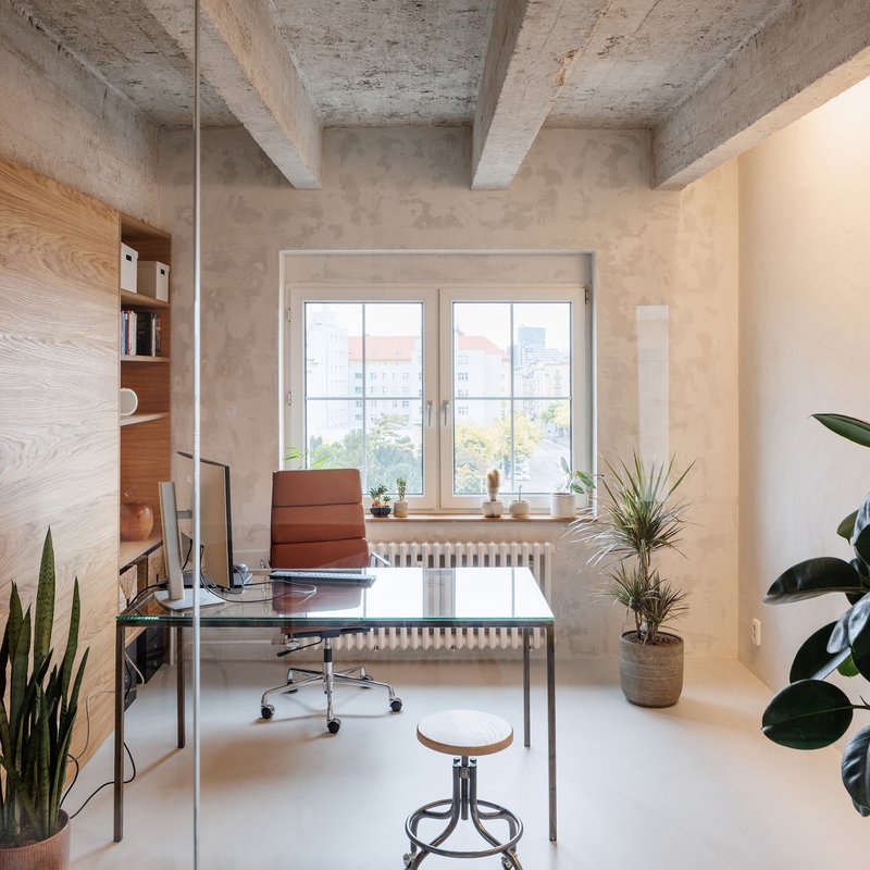 Office space with exposed concrete ceiling beams, glass desk and potted plants by the window