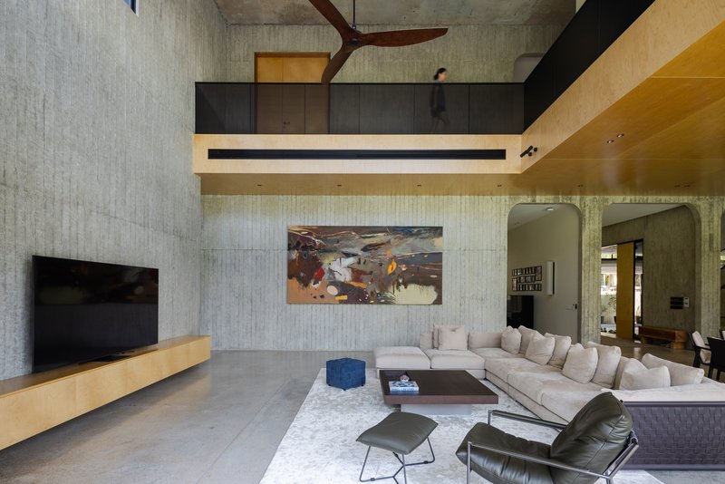 Double-height living room with board-formed concrete walls, exposed timber beams, and timber-lined mezzanine above