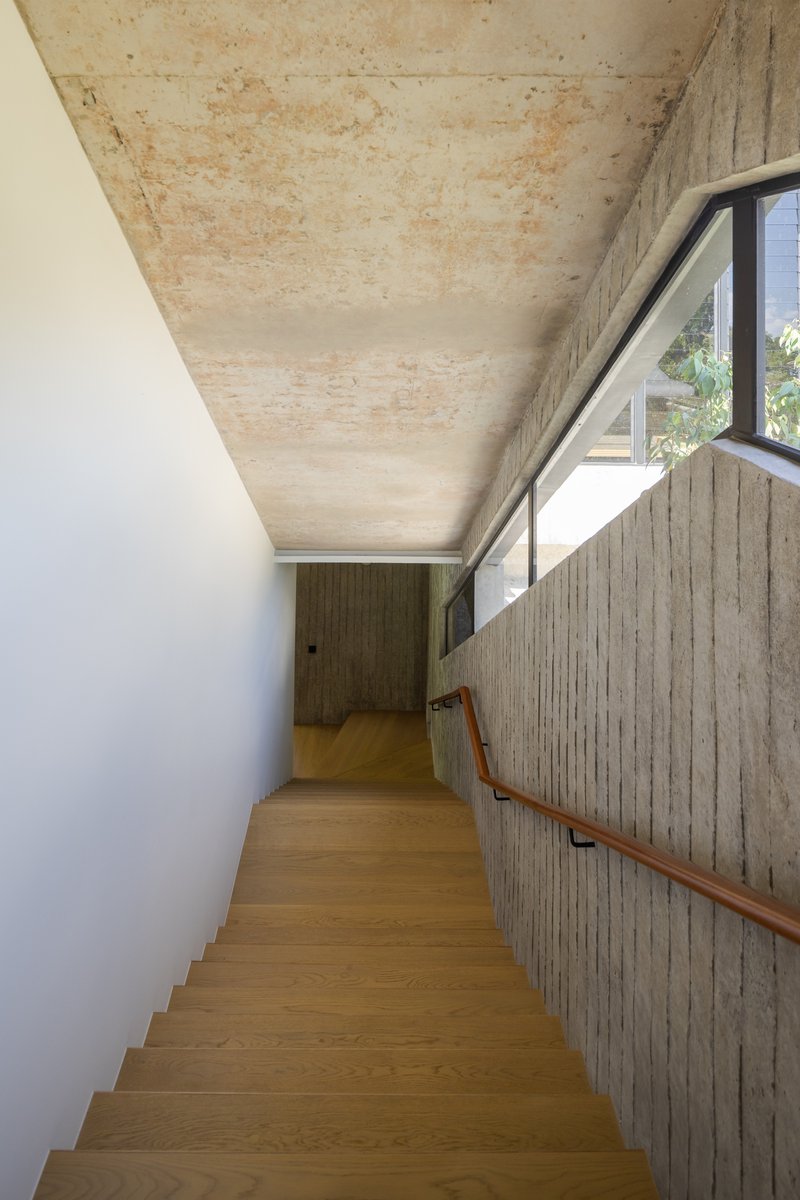 Timber stair ascending alongside board-formed concrete wall beneath an exposed plywood ceiling