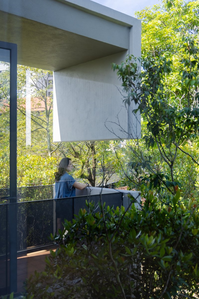 Covered balcony with person tending plants behind a dark metal railing amid dense foliage