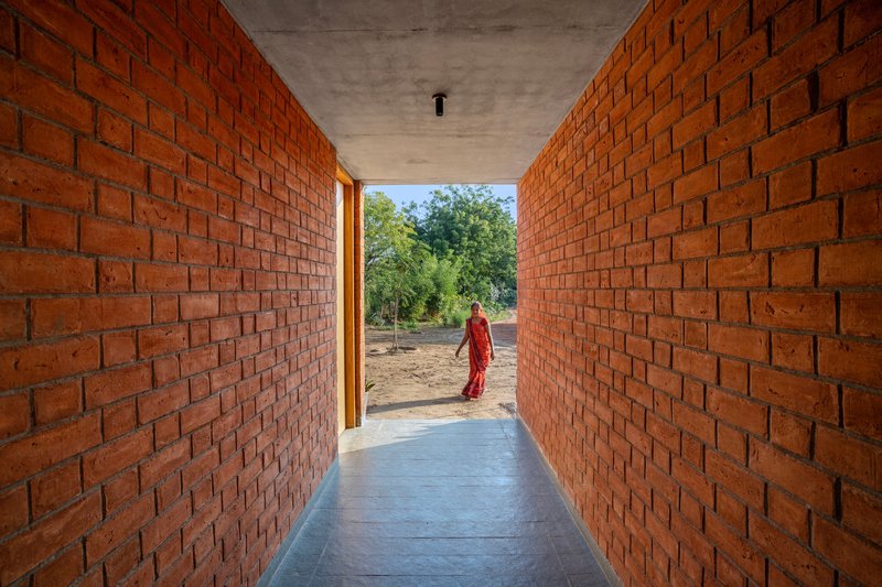 Corridor framed by exposed red brick walls leading to a figure in an orange sari