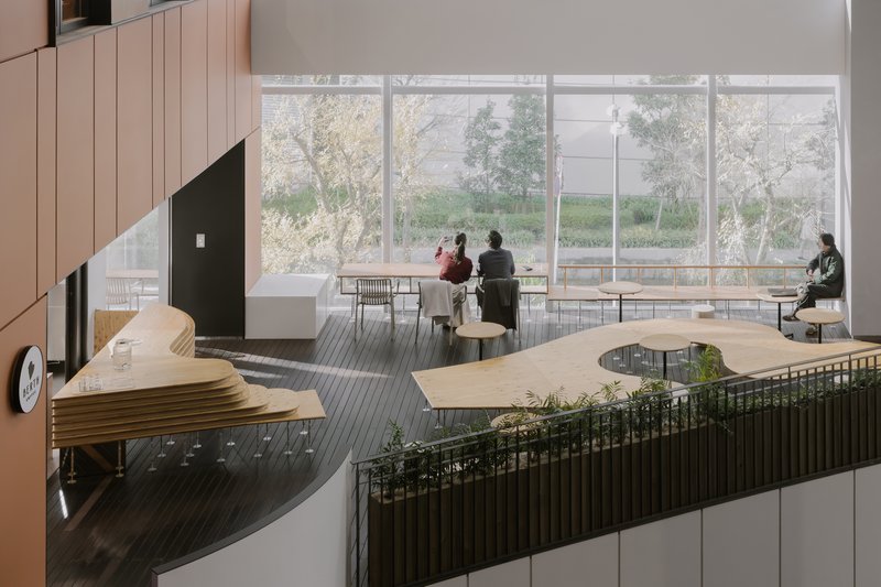 Double-height interior with organic plywood seating overlooking planted railings and floor-to-ceiling windows