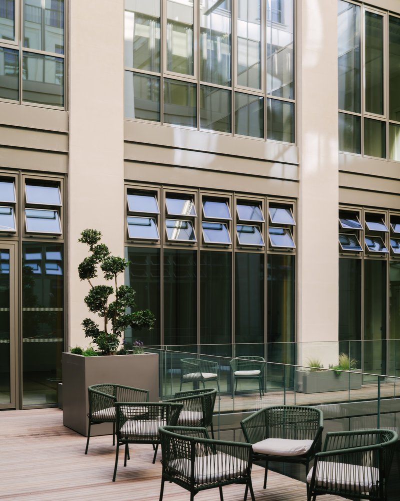 Courtyard terrace with louvered windows, glass balustrade, topiary tree in planter, and woven outdoor chairs