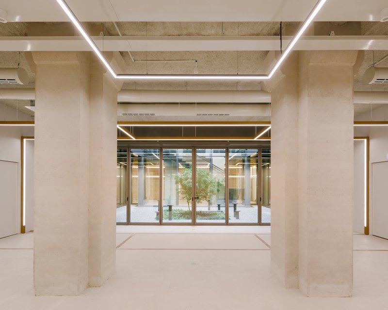 Interior space with exposed concrete ceiling beams and rectangular light fixtures framing a glazed courtyard entrance